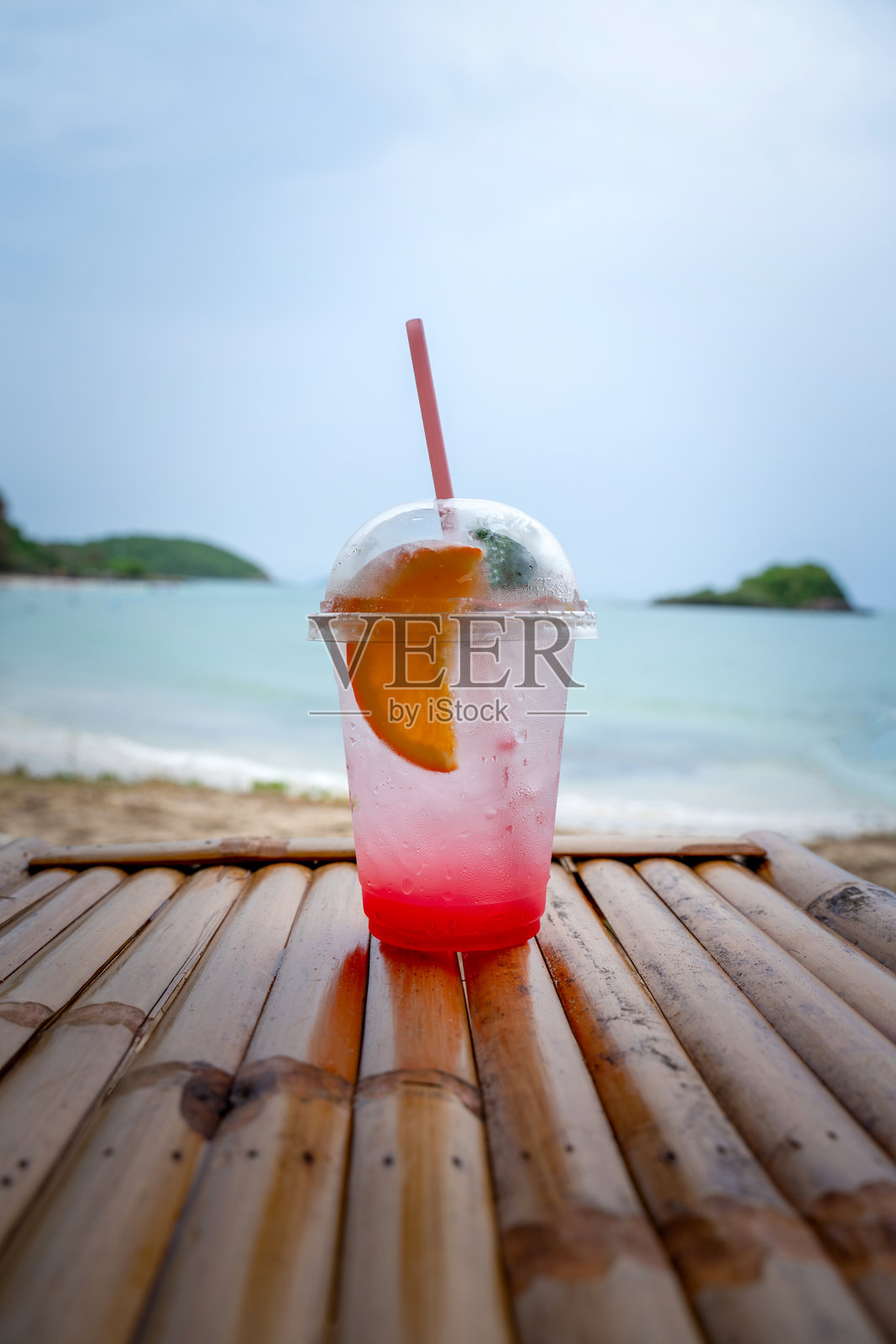 清爽的粉色柠檬水，配有橙片和吸管，放在塑料杯中，摆放在海滩的竹桌上，背景是热带海洋和岛屿。完美的夏日度假氛围。照片摄影图片