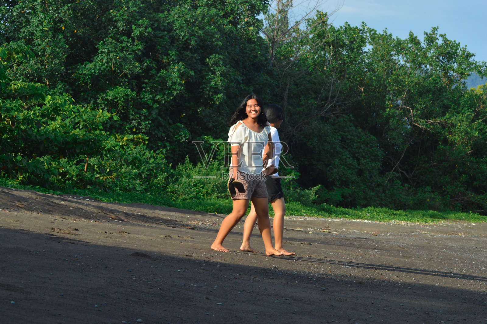两个赤脚的青少年在海滩上走着，手里拿着凉鞋。照片摄影图片