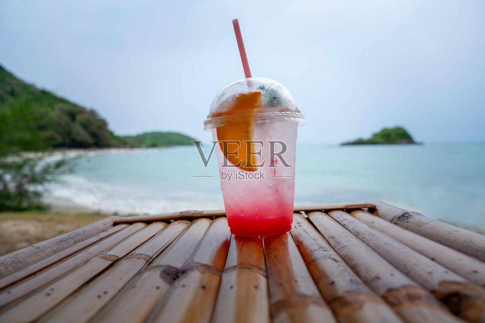 清新的粉色柠檬水，配有橙片和吸管，放在塑料杯中，摆放在海滩的竹桌上，背景是热带海洋和岛屿。完美的夏日度假氛围。照片摄影图片