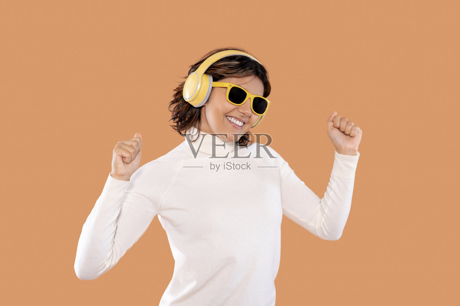 快乐的女性在棕色背景下戴着耳机和太阳镜跳舞，享受着阳光明媚的室内音乐。照片摄影图片