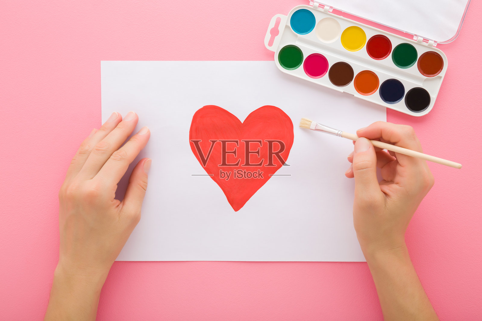 年轻女性手持画笔，在粉色桌面上用水彩在白纸上绘制红色心形。柔和的色调。特写镜头。爱情概念。俯视角度。照片摄影图片