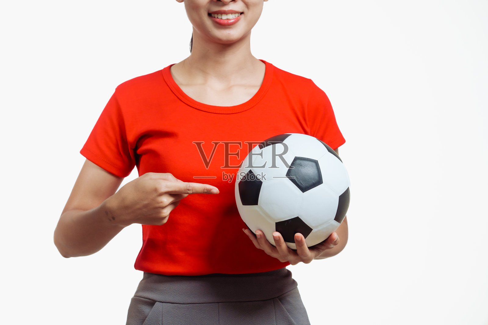 自信的年轻亚洲女性穿着鲜艳的红色衬衫，站在白色背景前，兴奋地为她最喜欢的足球队欢呼，手中握着足球，展现出热情的足球迷形象。照片摄影图片