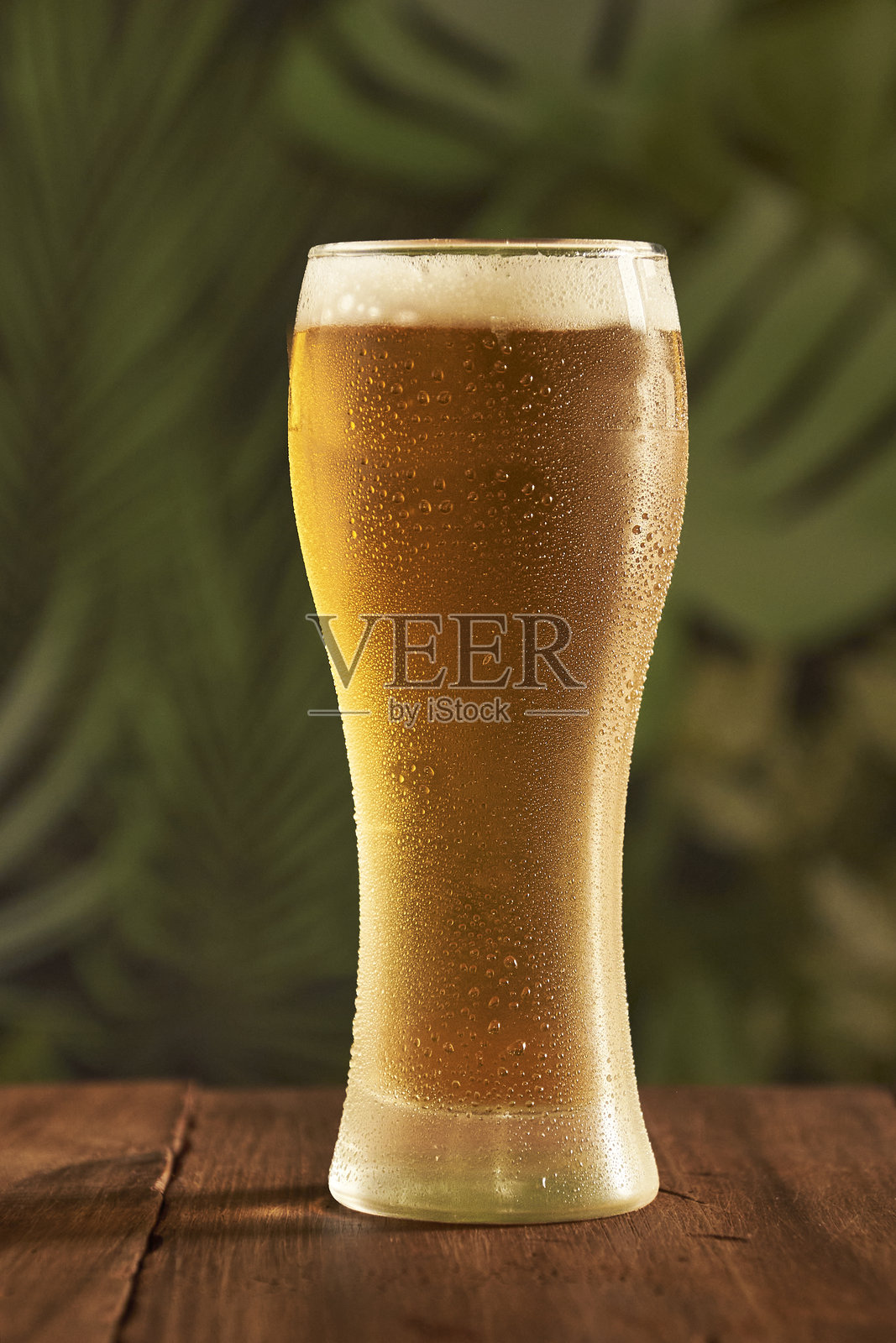 一杯冰冷的啤酒，前景有水滴，放在一个乡村风格的桌子上，背景是绿色的。照片摄影图片