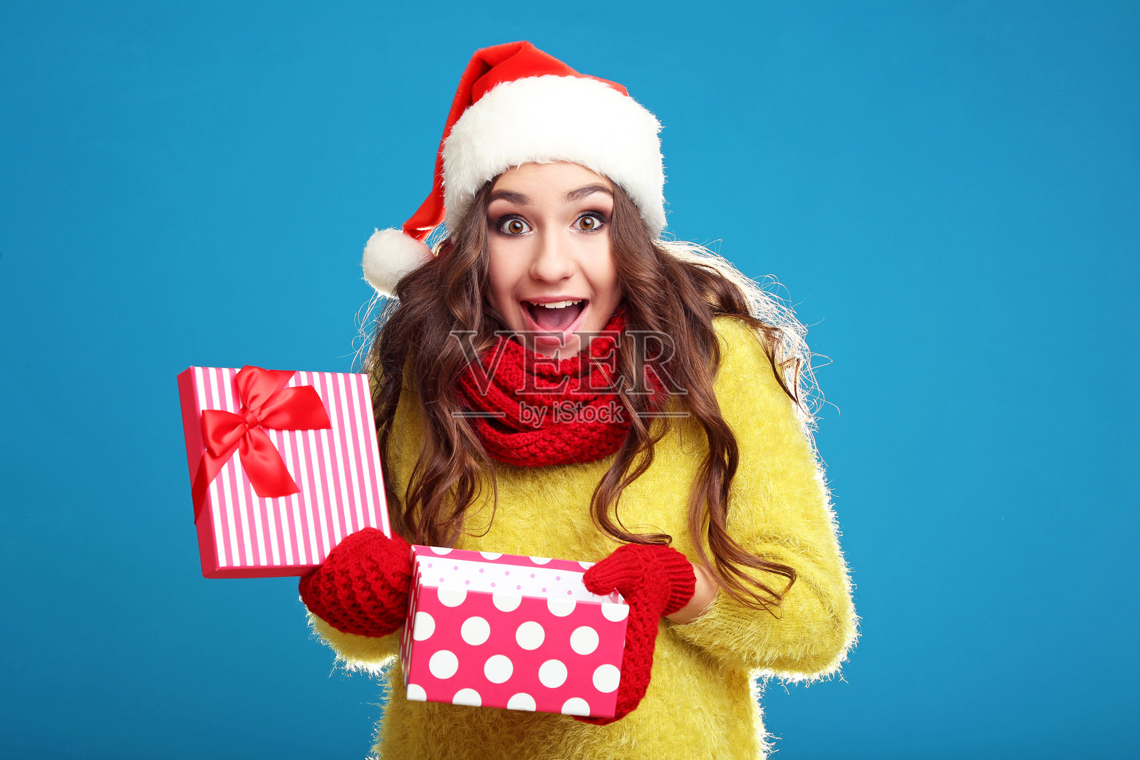 美丽的年轻女孩戴着圣诞帽，手里拿着礼物盒，背景为蓝色。照片摄影图片
