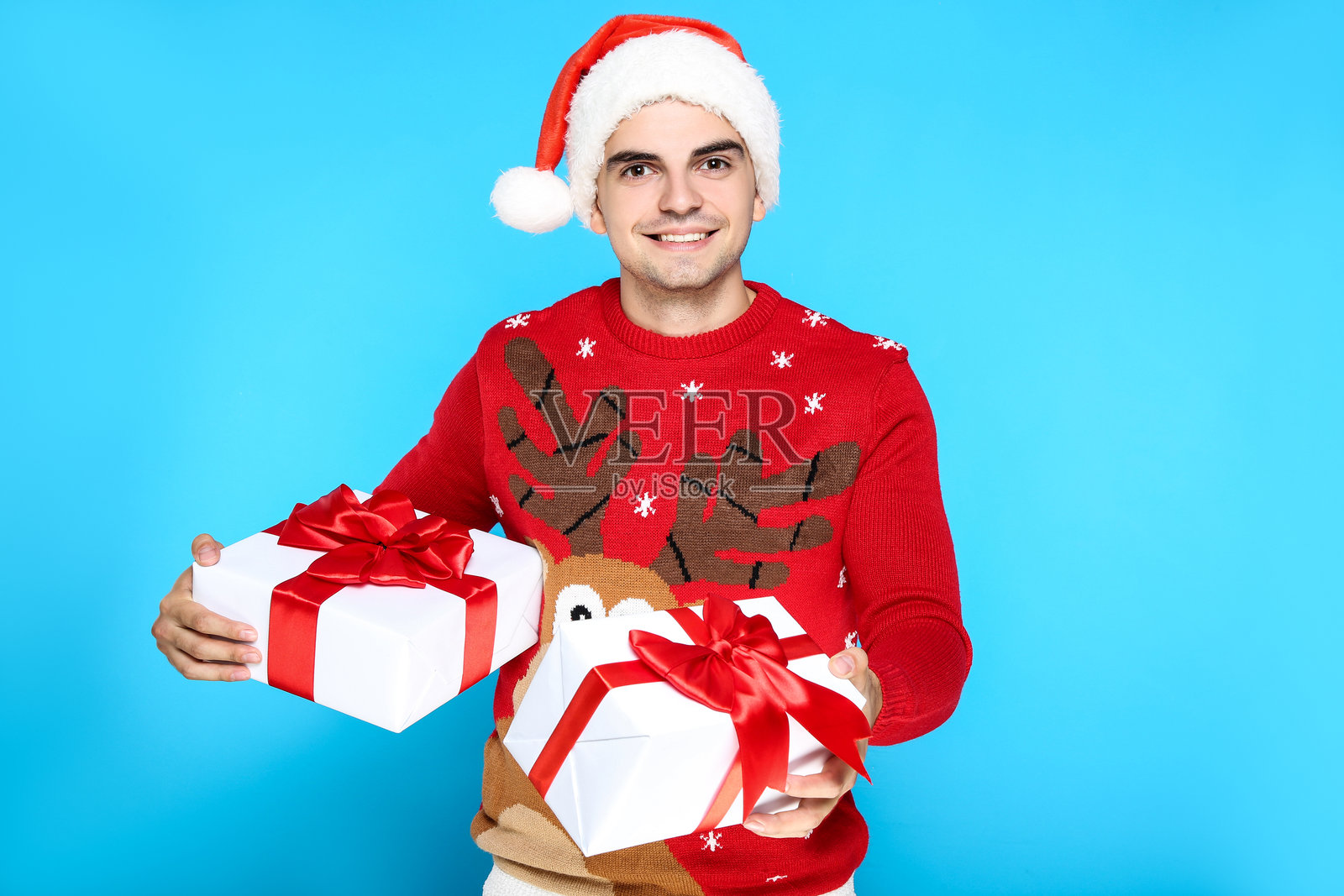 穿着圣诞毛衣、戴着圣诞老人帽、手持礼物盒的帅气男士，背景为蓝色。照片摄影图片
