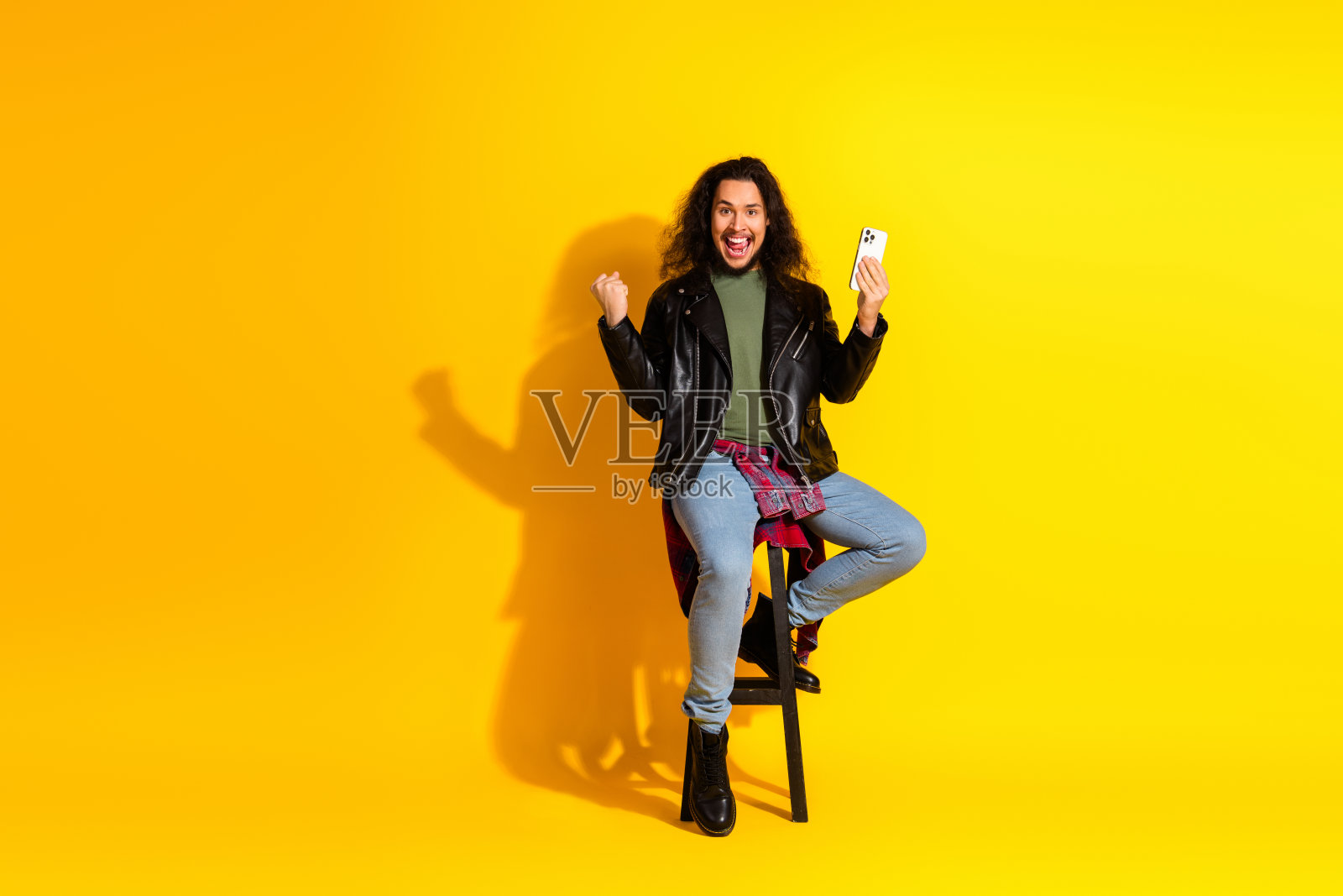 兴奋的年轻男子穿着黑色皮夹克，手持智能手机，背景为鲜艳的黄色，面露喜悦表情。照片摄影图片