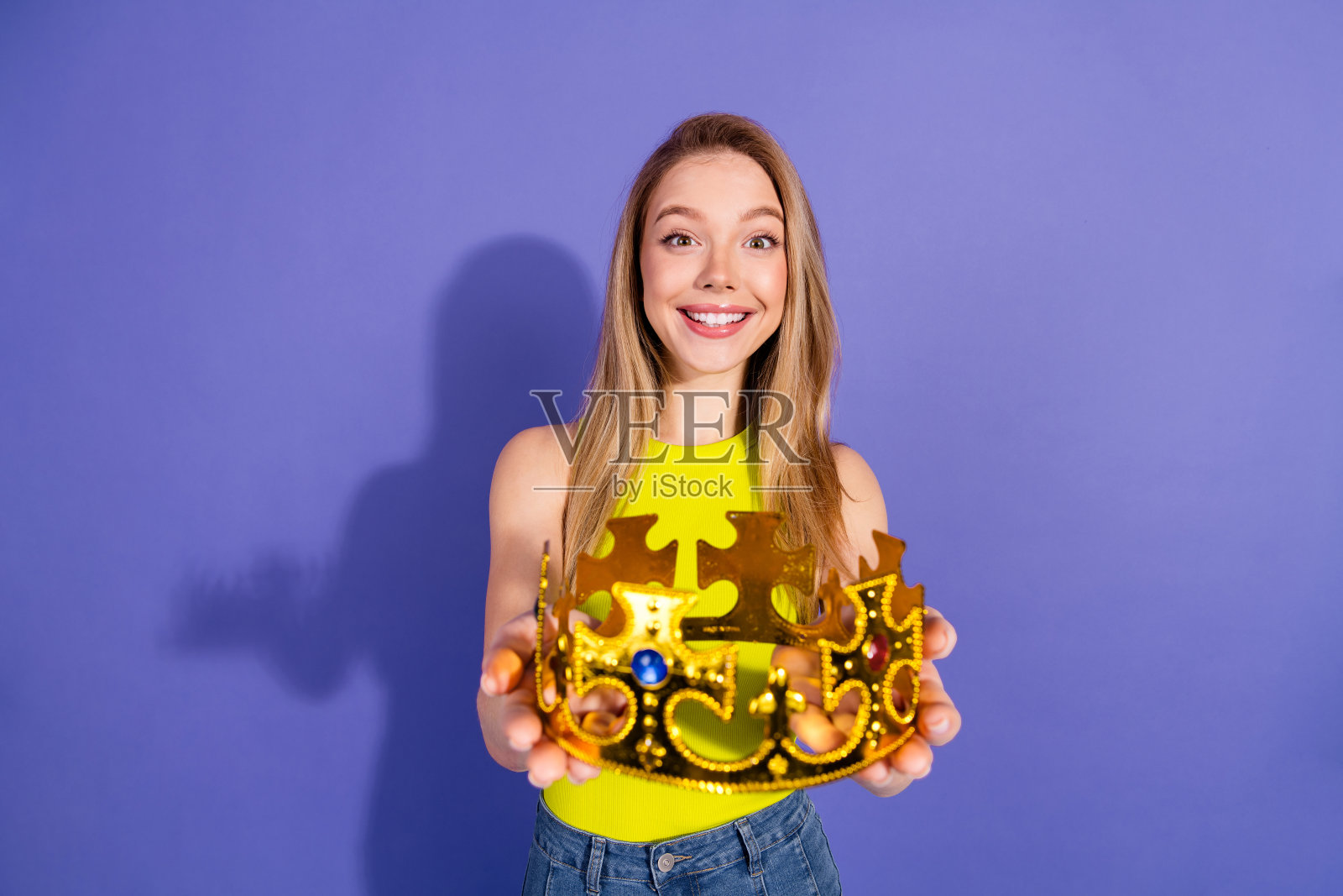 微笑的年轻女性双手捧着金色王冠，背景为紫色。照片摄影图片