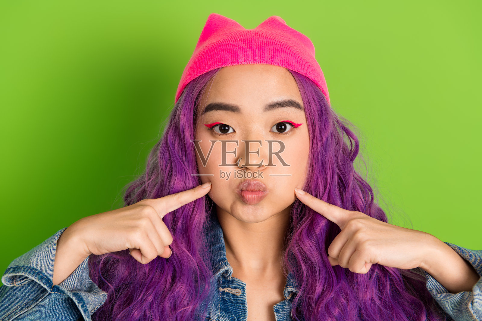 时尚的年轻女性，染着紫色头发，穿着粉色毛线帽和牛仔夹克，背景为绿色。照片摄影图片