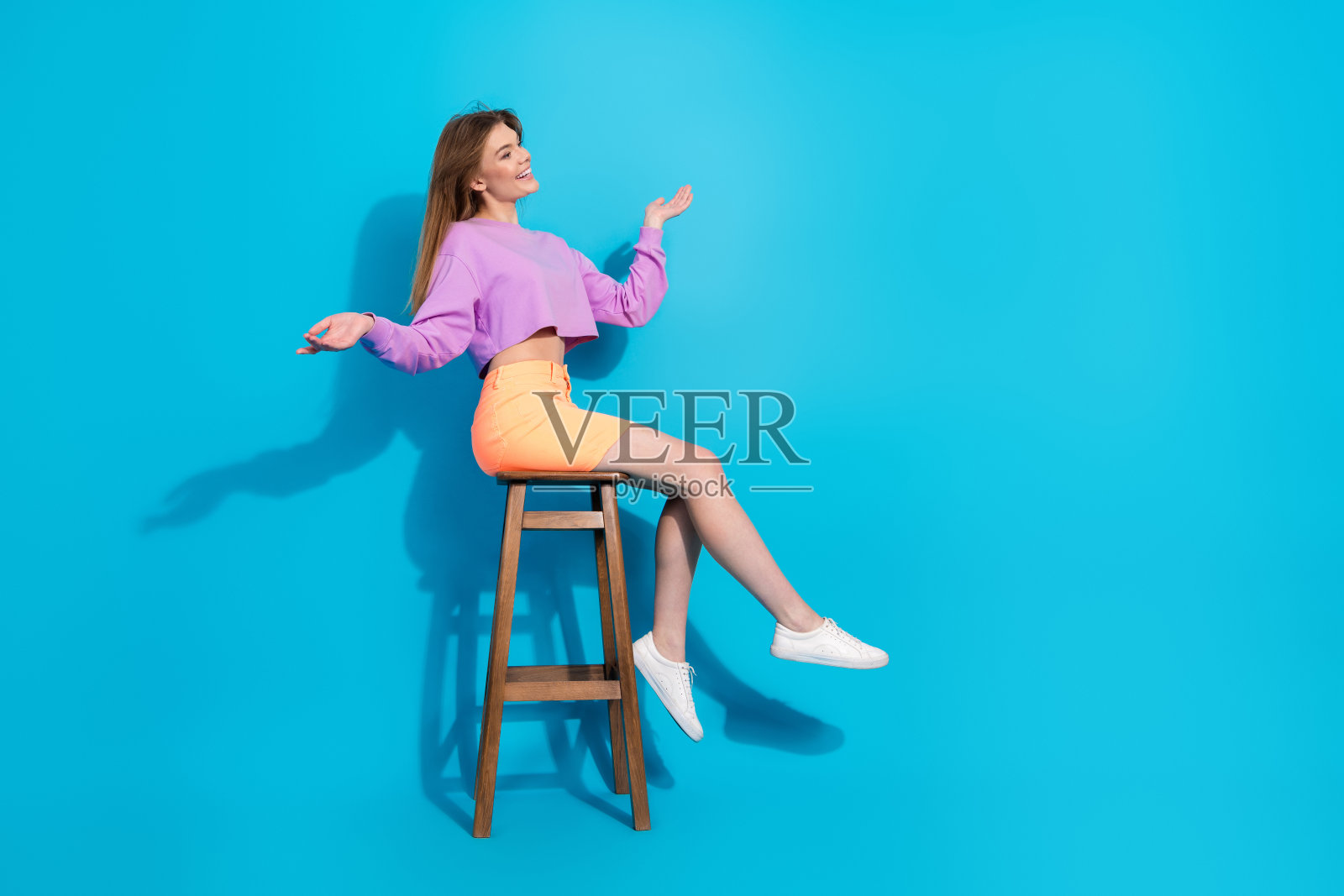 时尚女性穿着鲜艳的休闲风格，在明亮的蓝色背景前愉快地摆出姿势，坐在凳子上。照片摄影图片