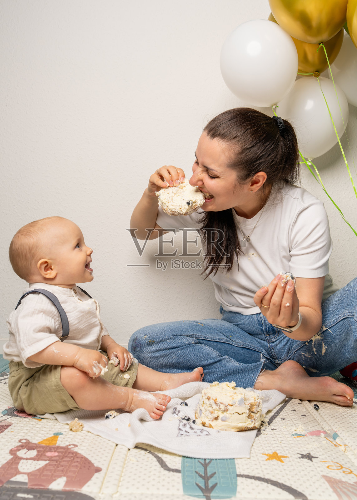 妈妈顽皮地咬了一口生日蛋糕，旁边一岁的小孩在开心地笑。背景是五彩气球。温馨的家庭时刻。母亲身份、节日、童年和家庭价值观的概念。照片摄影图片
