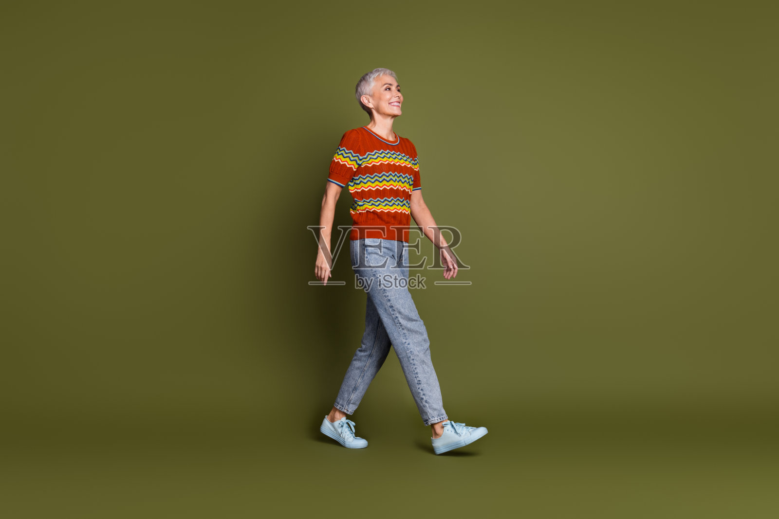 迷人的老年女性穿着时尚服装，自信地走在生动的卡其色背景前，展现出愉悦的表情。照片摄影图片