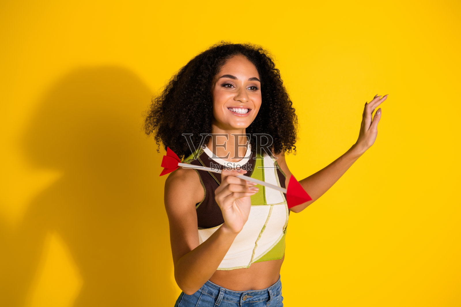 时尚的卷发女性穿着休闲上衣，手持箭矢，面带微笑，背景为鲜艳的黄色。照片摄影图片