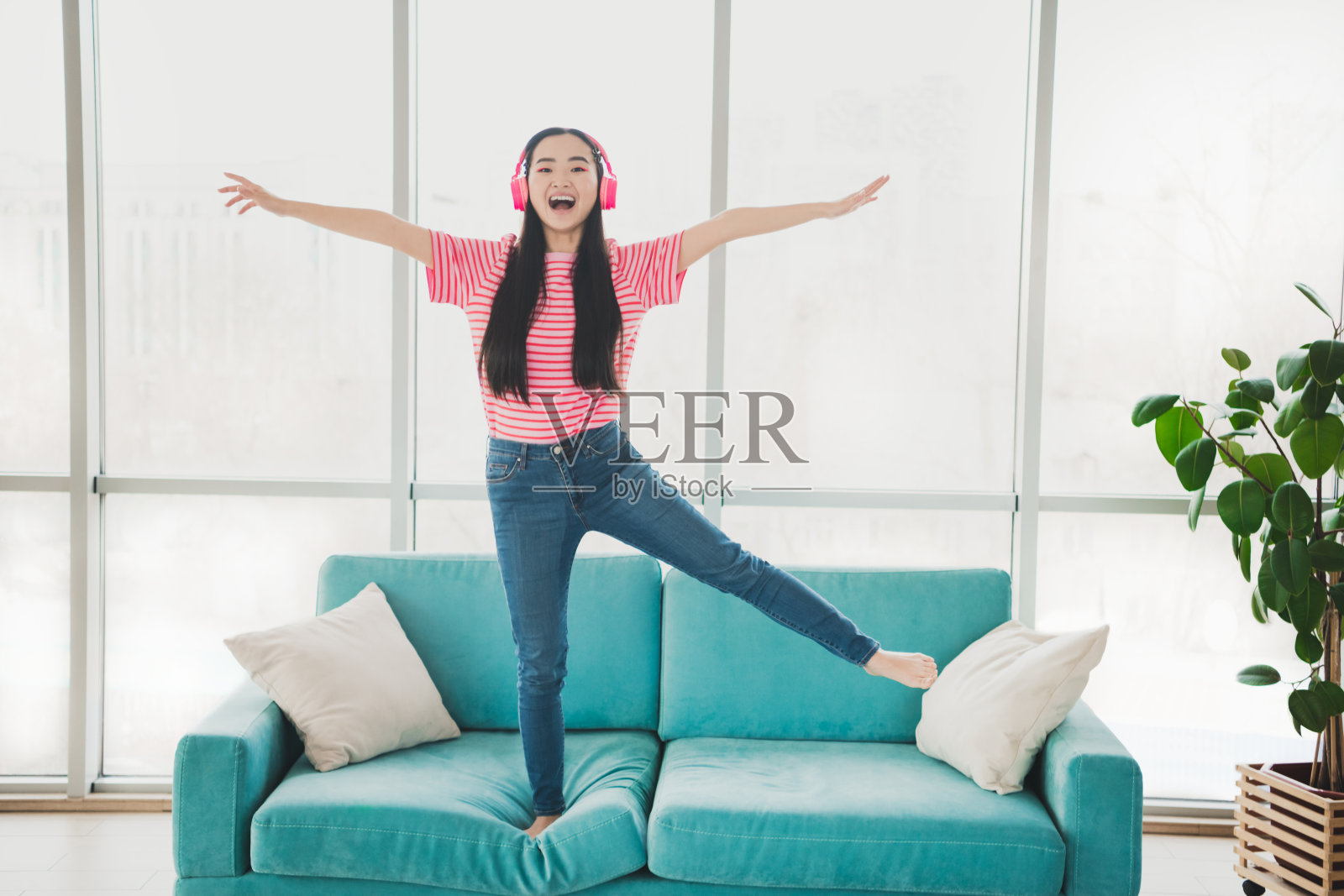 年轻女性在阳光明媚的舒适客厅里，戴着耳机快乐地在沙发上跳舞。照片摄影图片