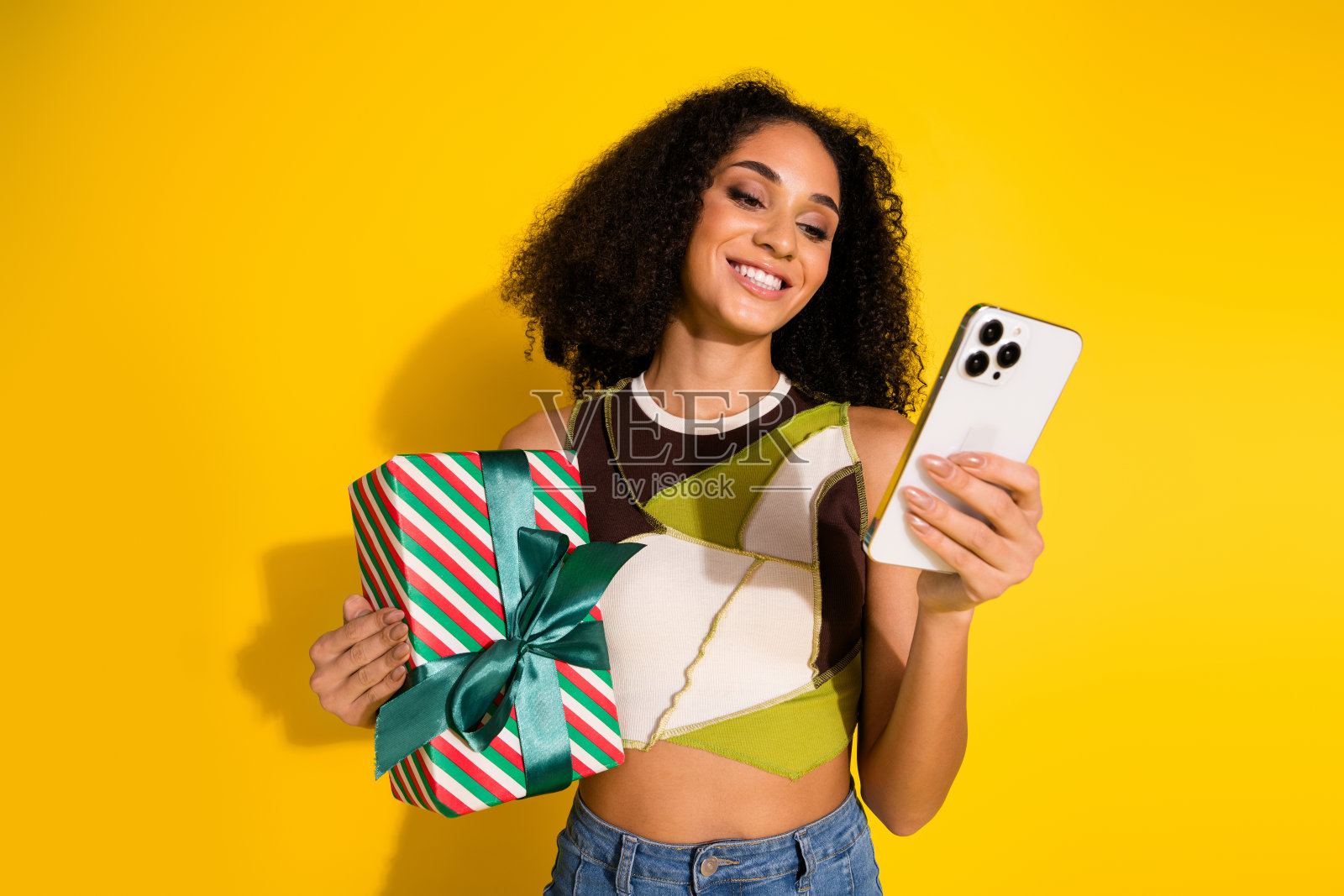 年轻女性手持礼物，凝视着智能手机，背景为鲜艳的黄色。照片摄影图片