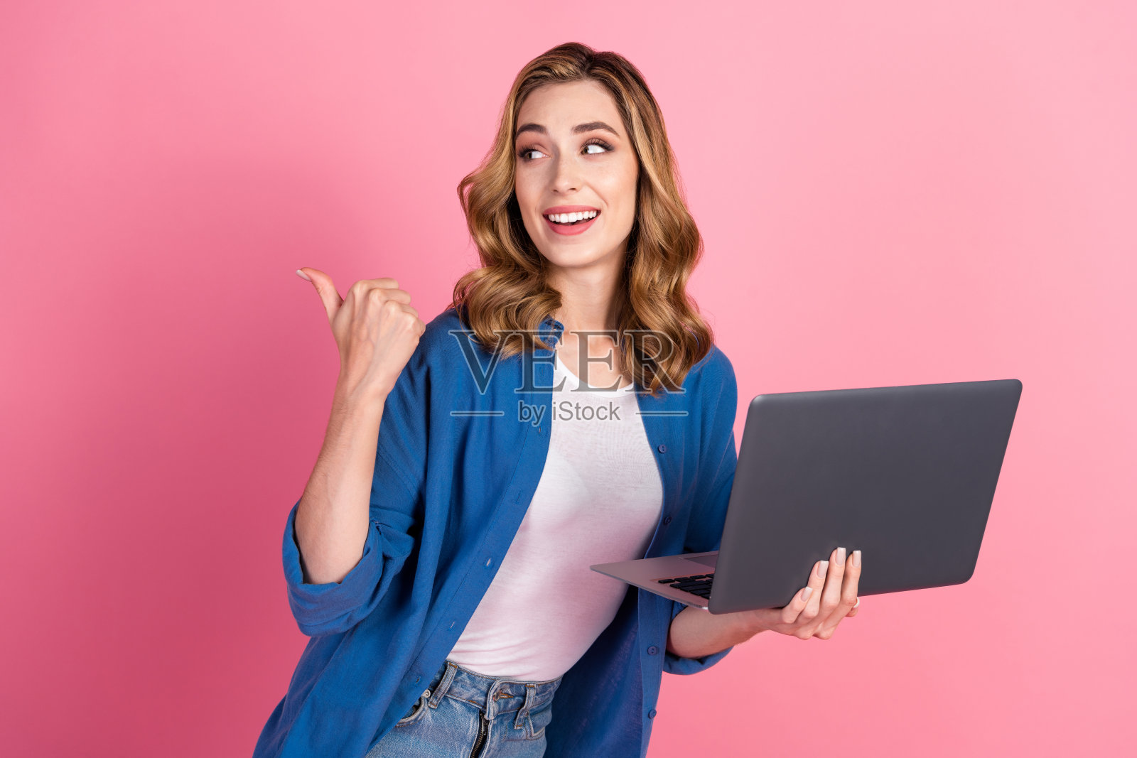 年轻女性穿着休闲蓝色衬衫，手持笔记本电脑并在粉色背景上做手势照片摄影图片