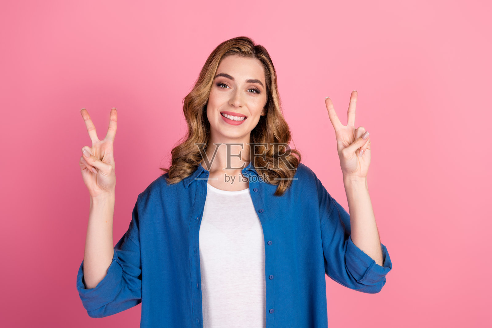 微笑的年轻女性穿着休闲蓝色衬衫，手做和平手势，背景为粉色。照片摄影图片
