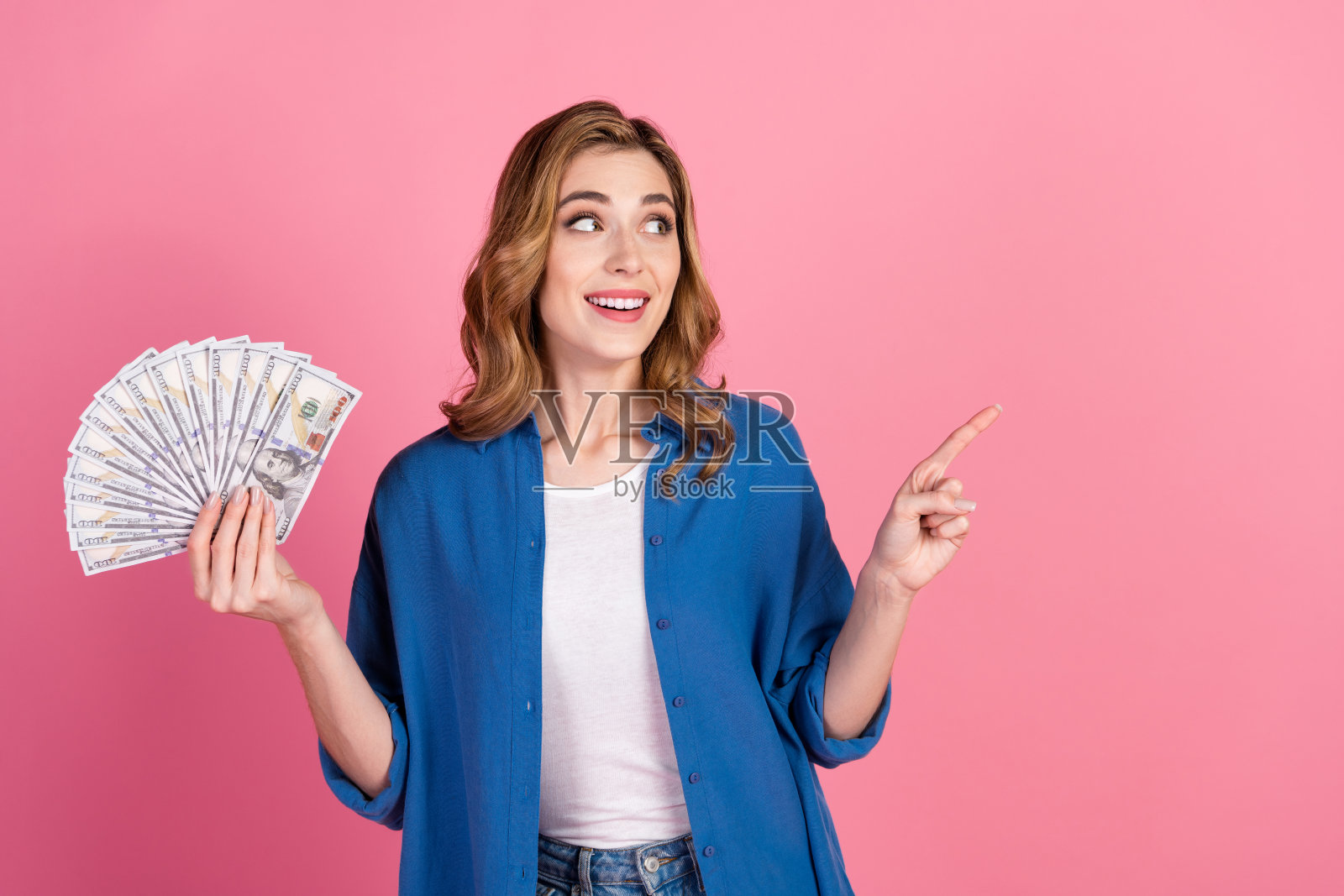 兴奋的年轻女性在粉色背景下手持现金并指向一侧照片摄影图片