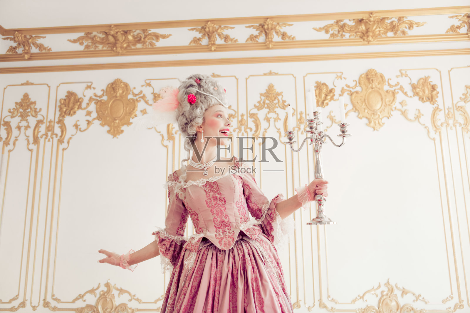一位年轻女性穿着优雅的粉色巴洛克长裙，站在奢华的宫殿内部，手中拿着一个银色烛台。照片摄影图片