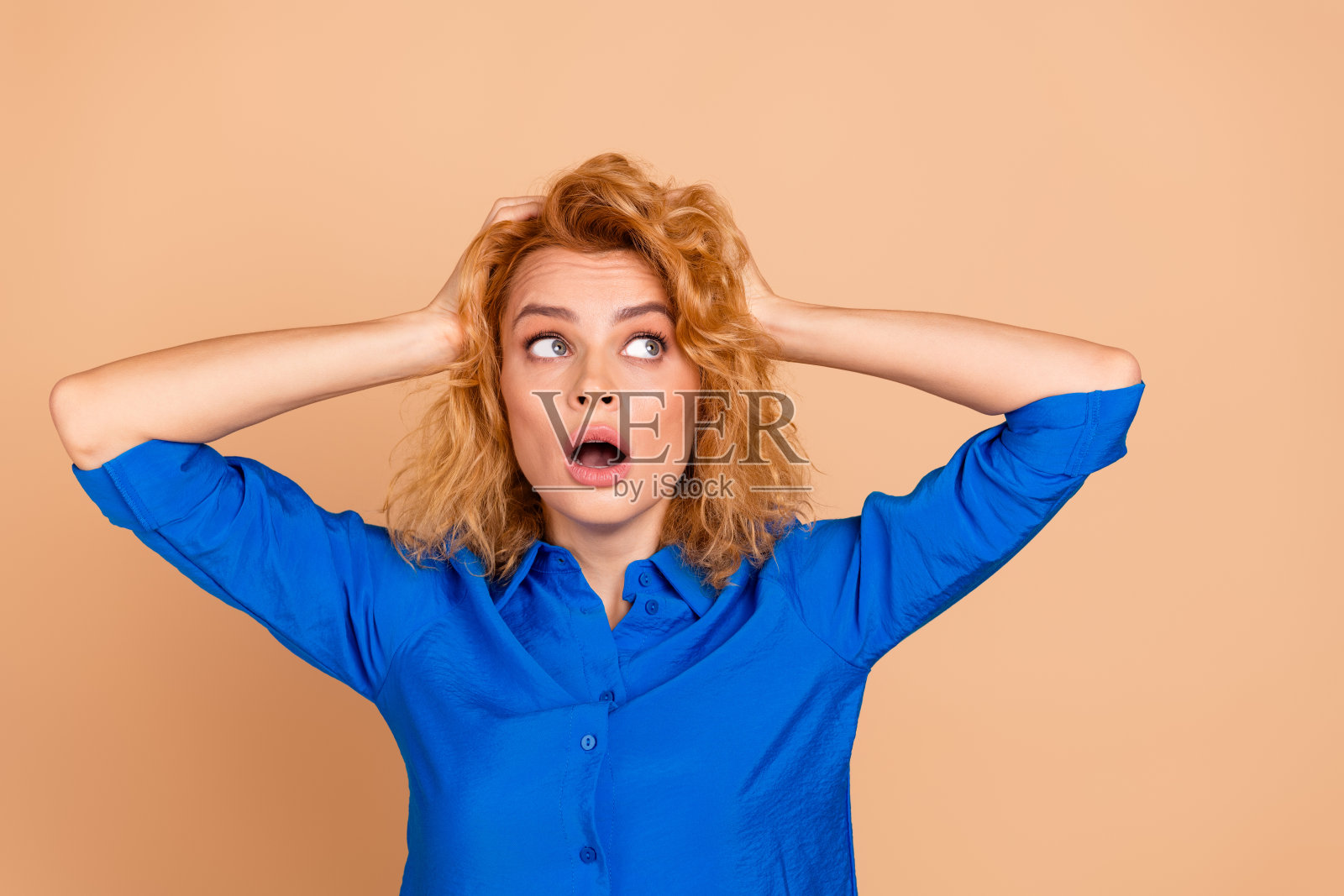 惊讶的年轻女性穿着休闲蓝色衬衫，双手放在头上，背景为米色。照片摄影图片