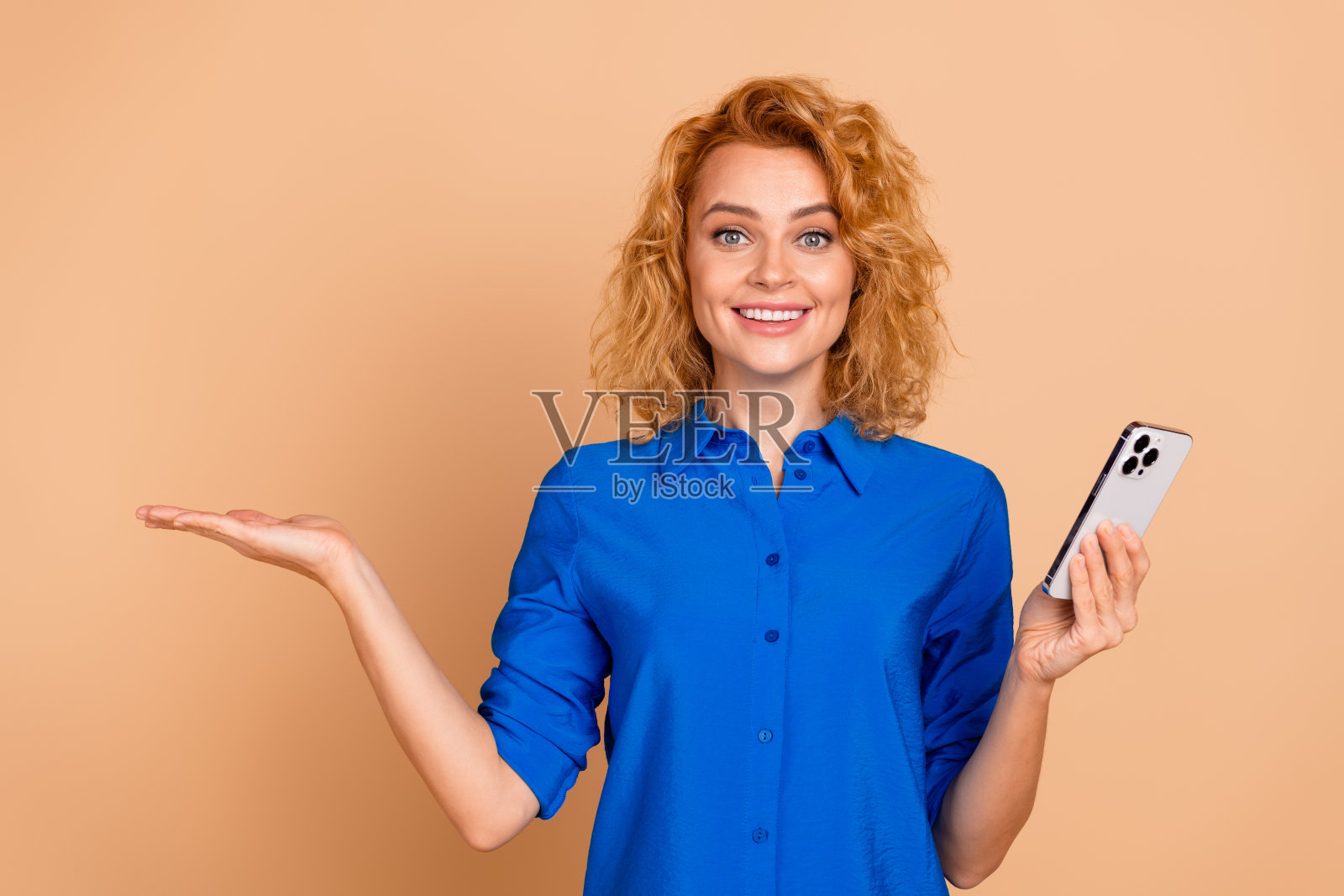 年轻女商人自信地微笑，展示智能手机，并在米色背景上伸出手进行演示。照片摄影图片