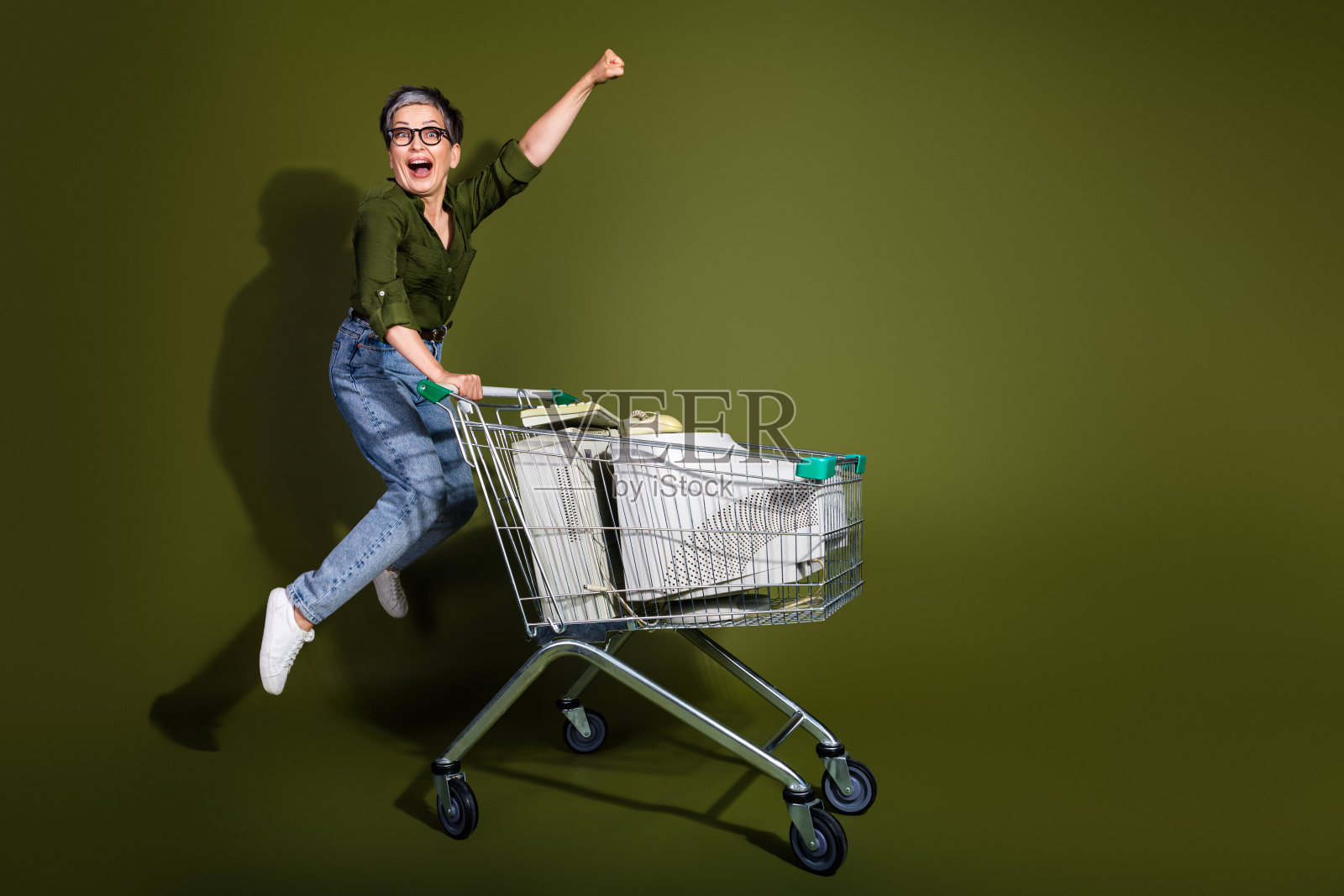 兴奋的成熟的女性在绿色背景下跳跃，展示出活力和快乐，旁边是装满物品的购物车。照片摄影图片