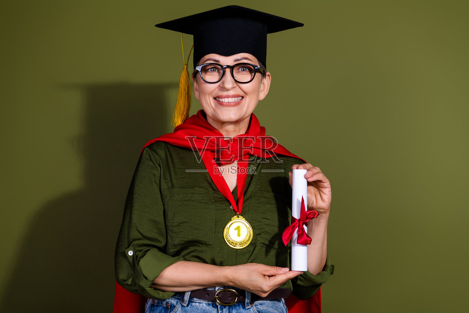 微笑的成熟的女性戴着毕业帽，手持文凭，在绿色背景下庆祝成就，佩戴奖牌，穿着休闲服装。照片摄影图片