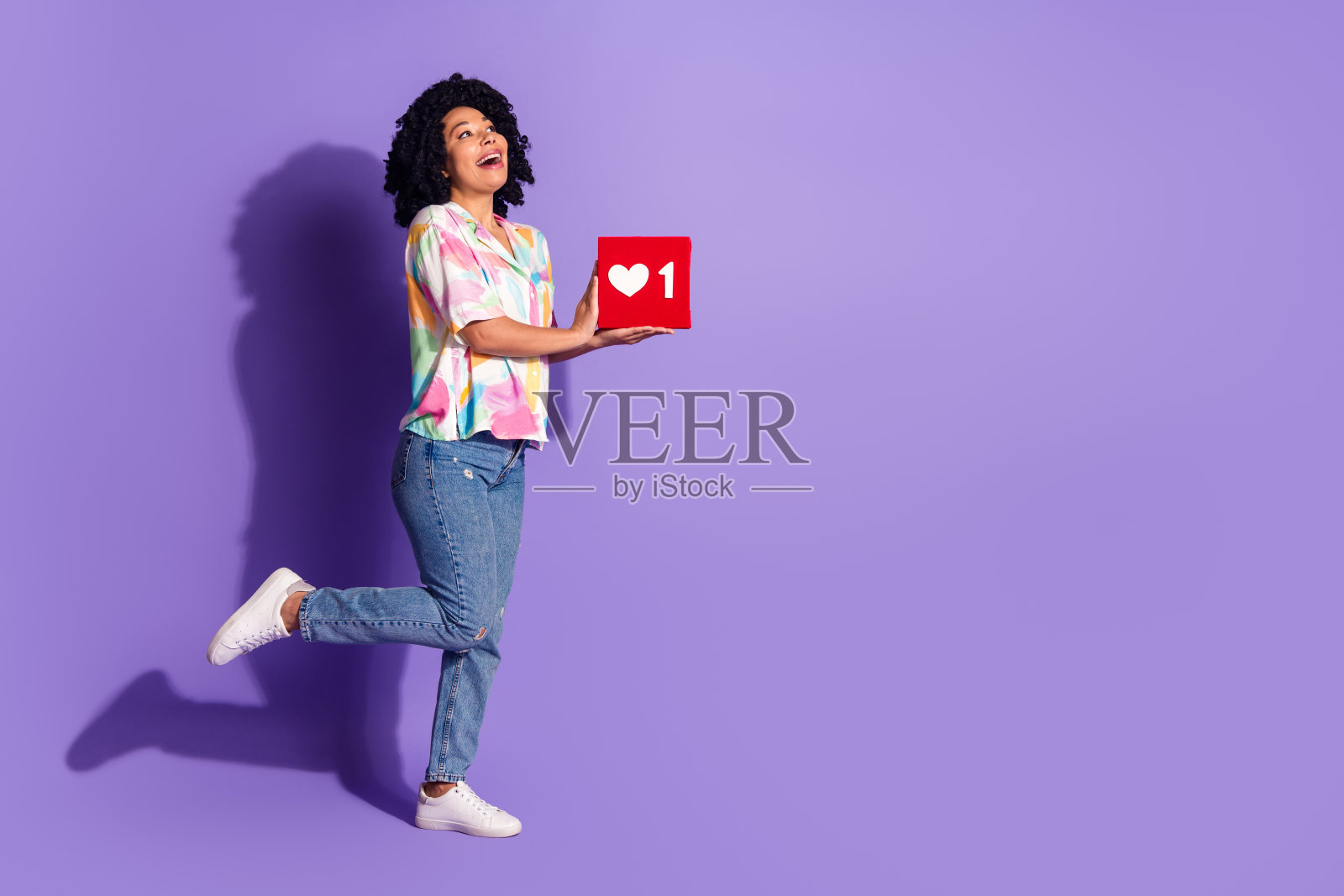 年轻女性穿着休闲多彩的服装，手持一个带有心形符号的红色立方体，背景为紫色，表达出快乐和兴奋。照片摄影图片