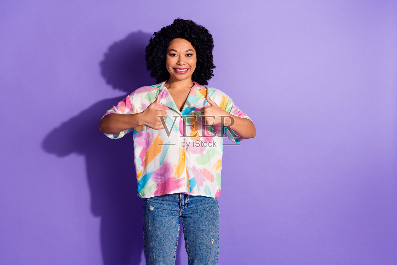 微笑的年轻女性在鲜艳的紫色背景下竖起大拇指，穿着休闲的多彩服装照片摄影图片