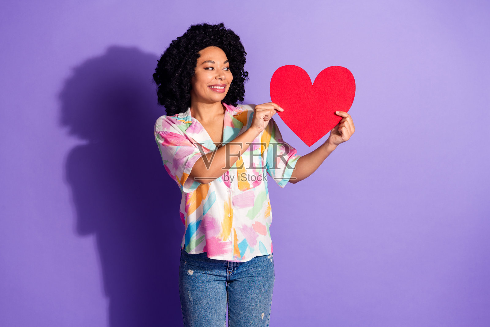 年轻的非裔美国女性在紫色背景下做出心形手势，穿着色彩鲜艳的衬衫，面带愉悦的表情，展示时尚。照片摄影图片