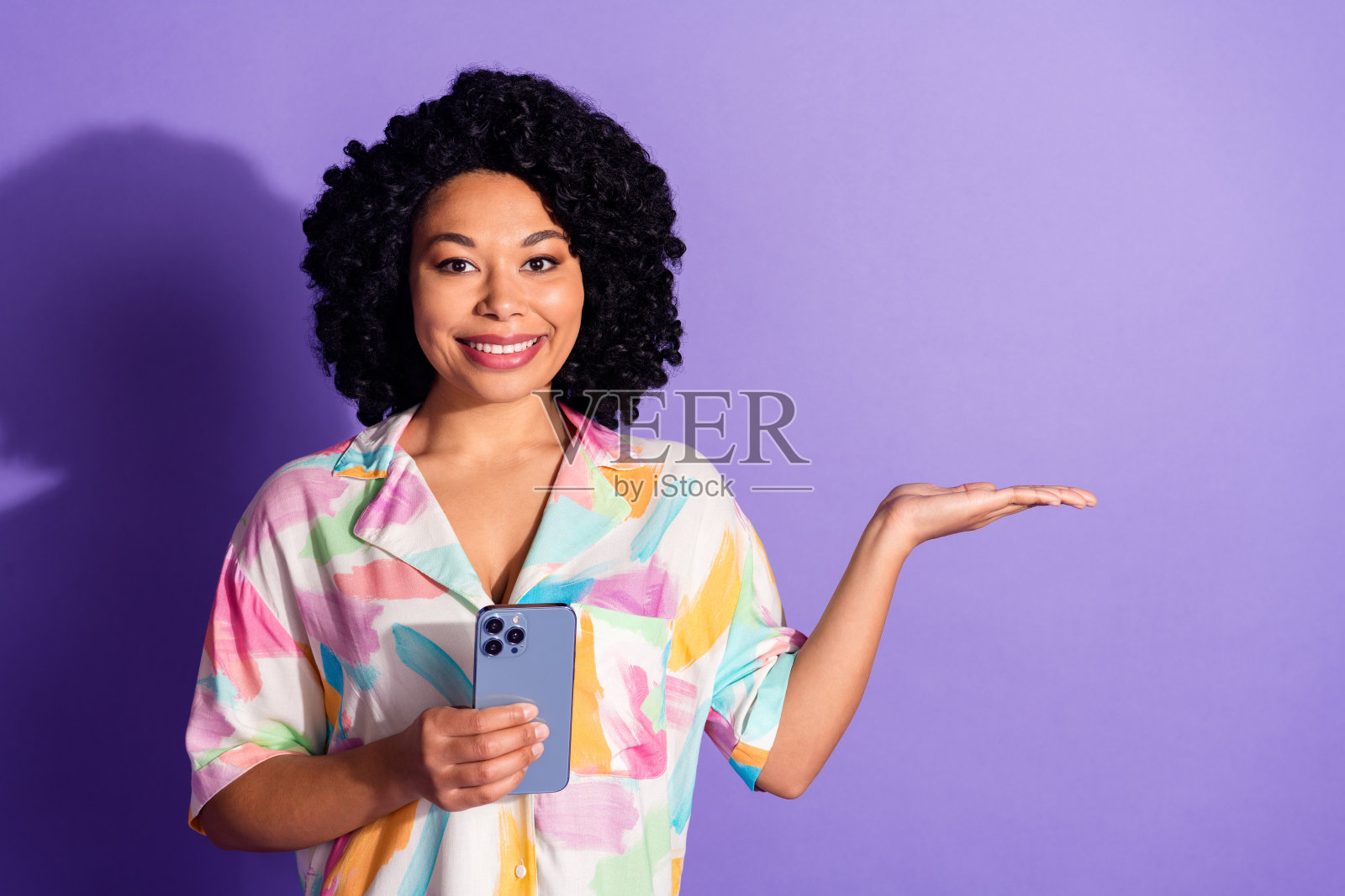 年轻女性卷发，手持手机并做手势，背景色彩丰富，展示休闲时尚的夏季服装和生活方式。照片摄影图片