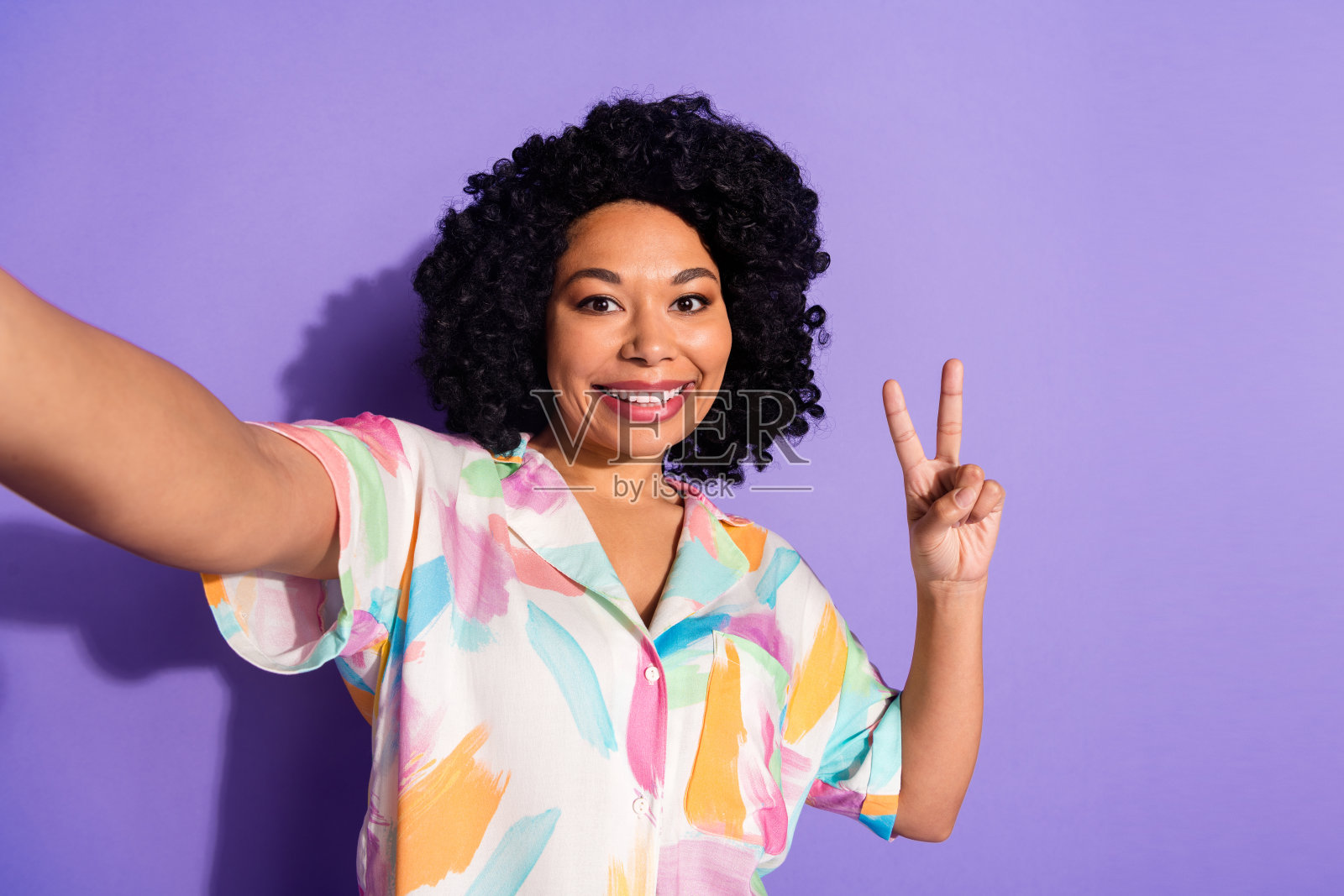 自信的年轻女性在紫色背景前摆姿势，微笑着比出和平手势，穿着色彩鲜艳的夏季衬衫。照片摄影图片