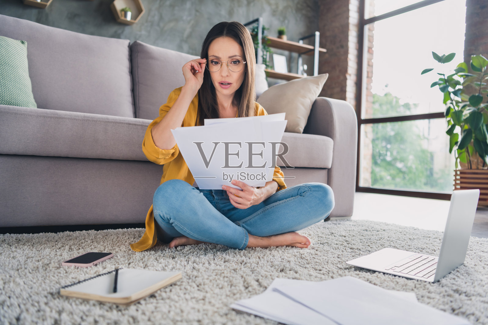 一位穿着黄色衬衫的年轻女士在室内家中认真忙碌，使用现代设备阅读文件。照片摄影图片