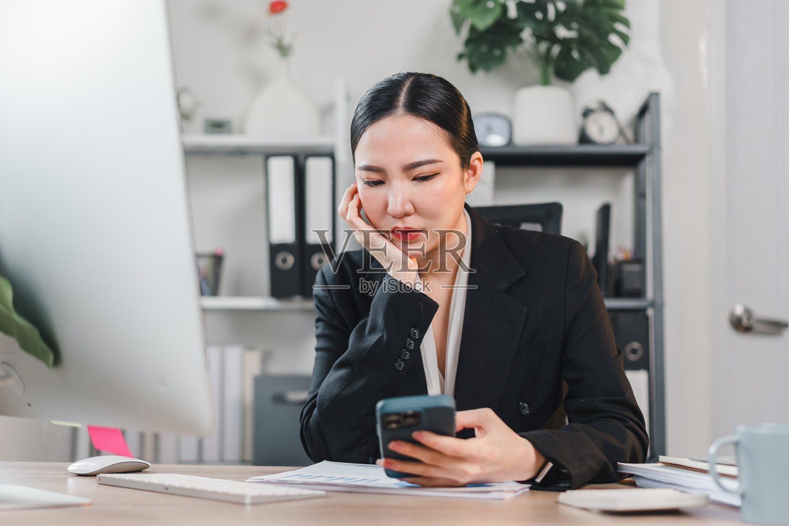 专注的亚洲女商人在现代办公室环境中穿着黑色西装坐在桌子旁，查看她的智能手机。照片摄影图片