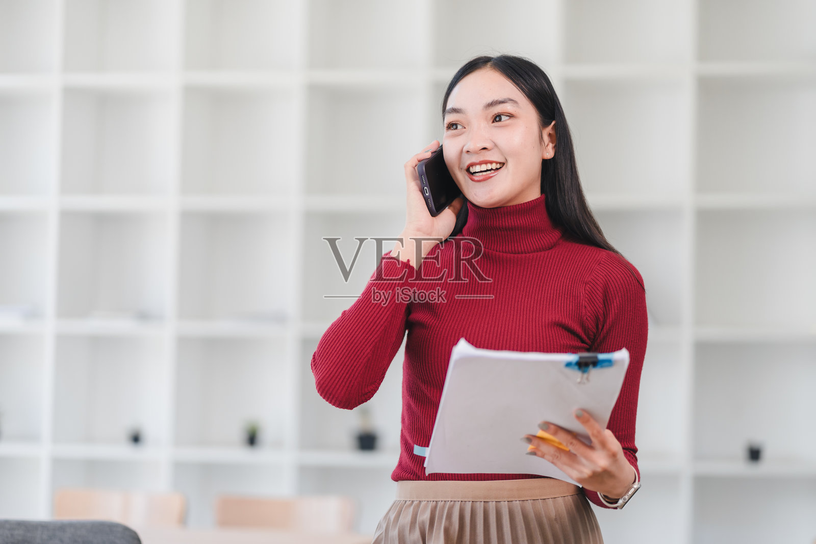 穿着红色高领毛衣的快乐亚洲女性在现代办公室里一边打电话一边拿着文件。照片摄影图片