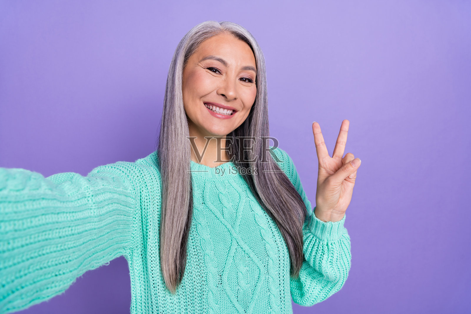 快乐的成熟的女性在紫色背景前自拍，做出和平的V字手势照片摄影图片