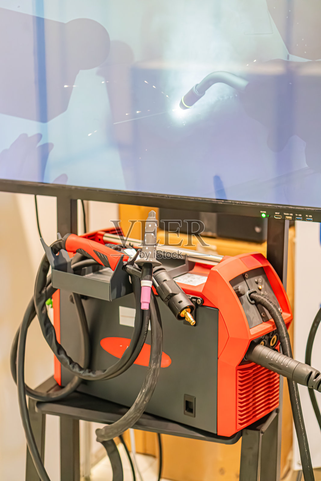 在模糊的车间环境中，一款红色的电池驱动工业工具，可能是钻磨机，部分被管道或风管遮挡。该工具正在运行，灯光和电线可见。照片摄影图片