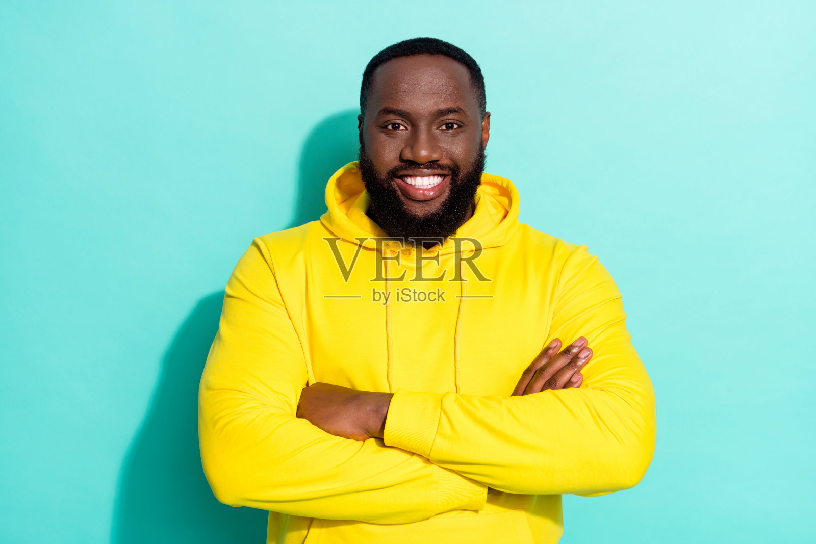 穿着黄色运动服的棕发酷炫千禧一代男士，双臂交叉，背景为青色。照片摄影图片
