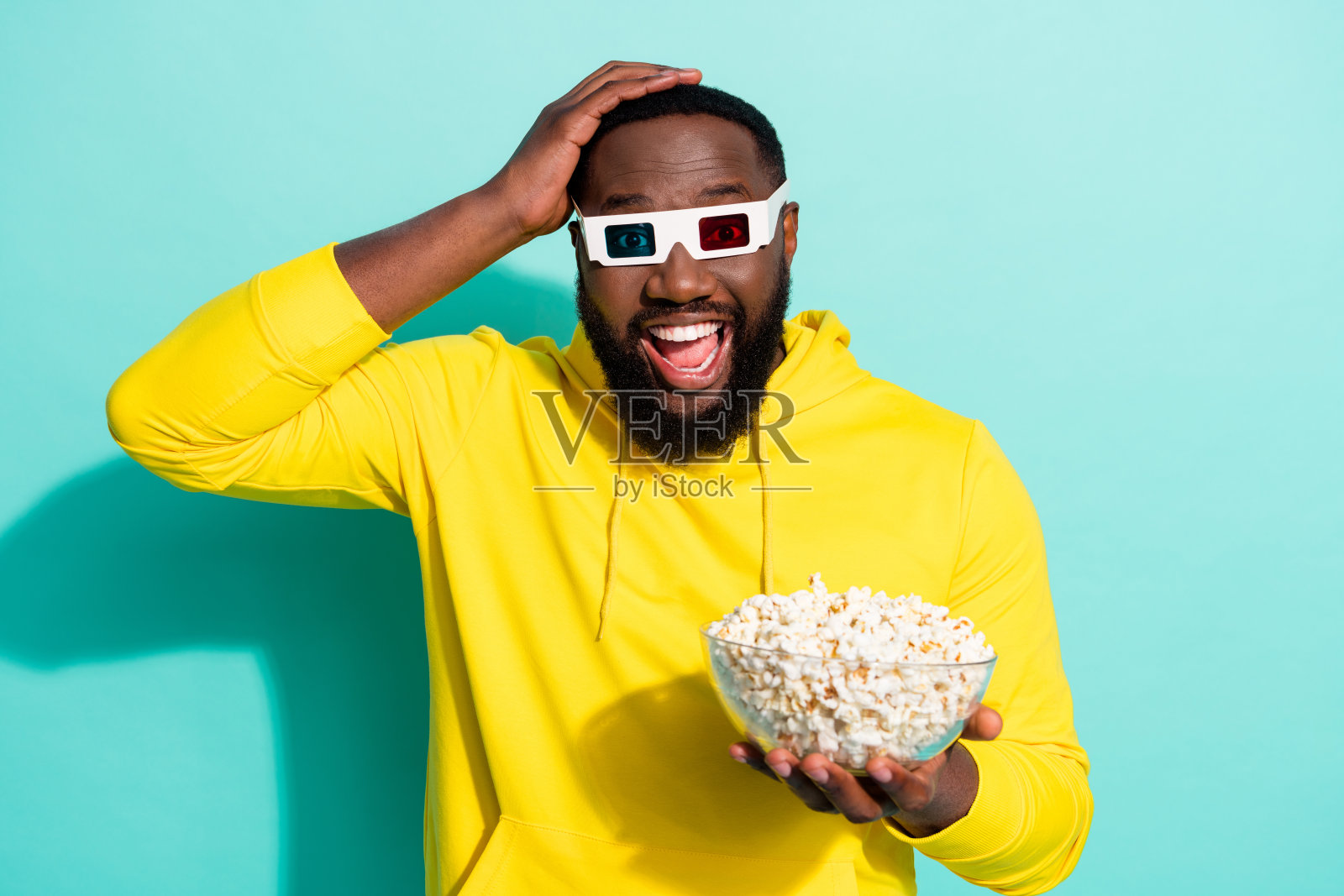 惊讶的男孩在青色背景下戴着3D眼镜，手里拿着爆米花盘，观看影院大片。照片摄影图片