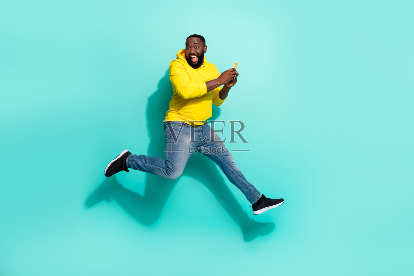 全身照片，疯狂的男人在青色背景下快速跳跃奔跑，使用智能手机讨价还价。照片摄影图片