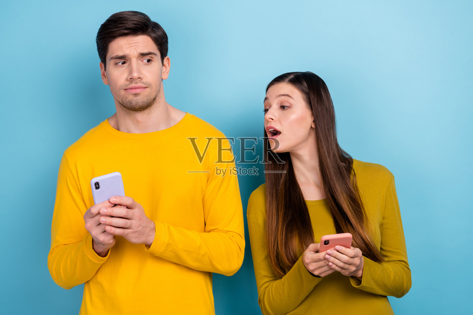 好奇的女士看着丈夫的手机屏幕，不想让她看到他的社交媒体动态，背景为蓝色。照片摄影图片