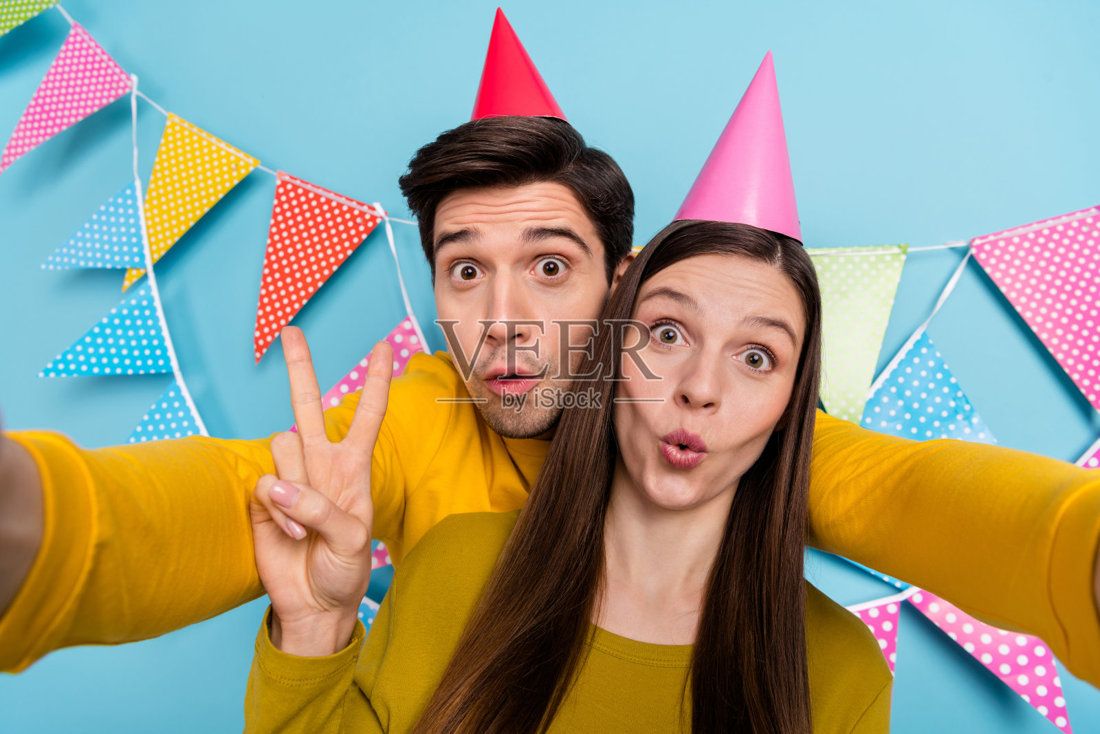 惊讶的已婚男士与女士自拍，做出V字手势，视频通话，节日场合，背景为蓝色。照片摄影图片