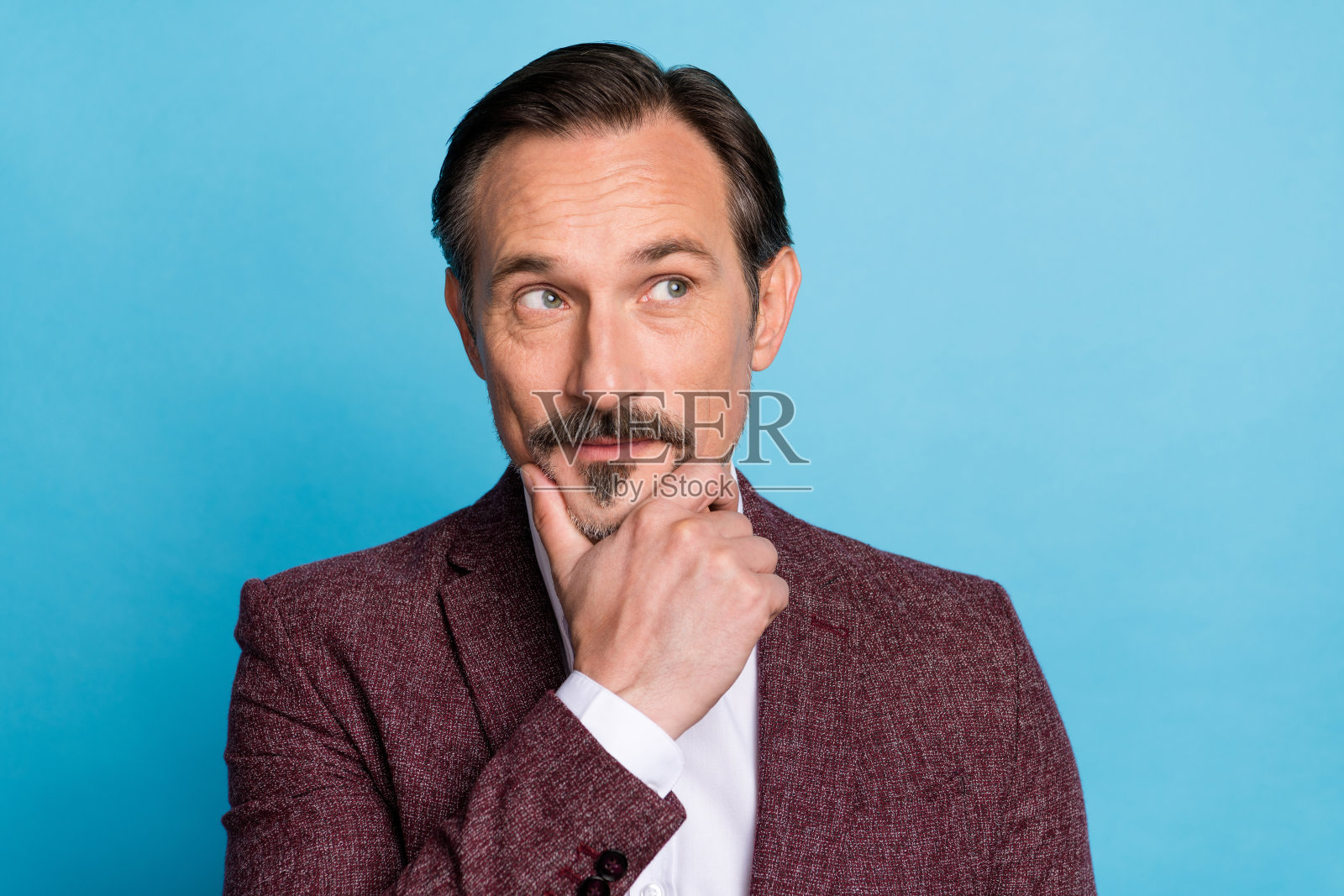 聪明谨慎的胡子男人凝视空旷的空间，寻找解决方案，穿着红色西装外套，背景为蓝色。照片摄影图片
