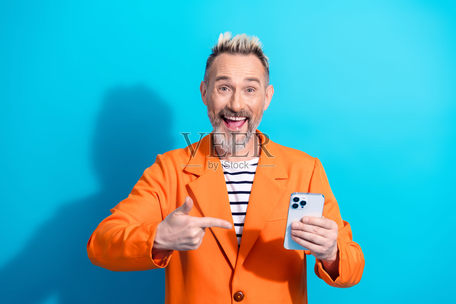 充满魅力的成熟男性穿着鲜艳的橙色夹克，展示着智能手机，背景是明亮的蓝色，散发出自信的气息。照片摄影图片