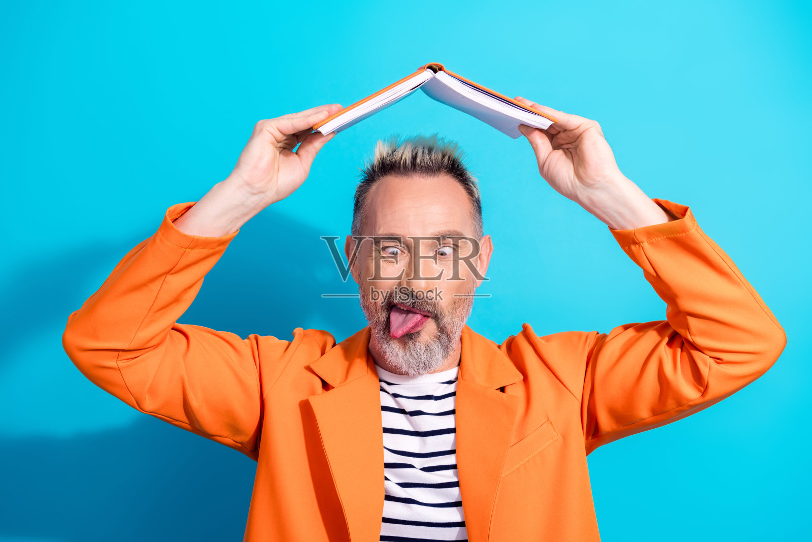 充满魅力的成熟男士在鲜艳的蓝色背景下，穿着橙色西装，开心地把书当作帽子。照片摄影图片