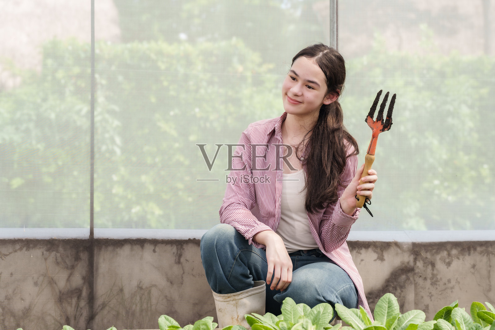 亚洲女学生坐在学校温室里微笑，手里拿着小耙子，刚刚参加完农业学习课程中的动手蔬菜种植活动。照片摄影图片