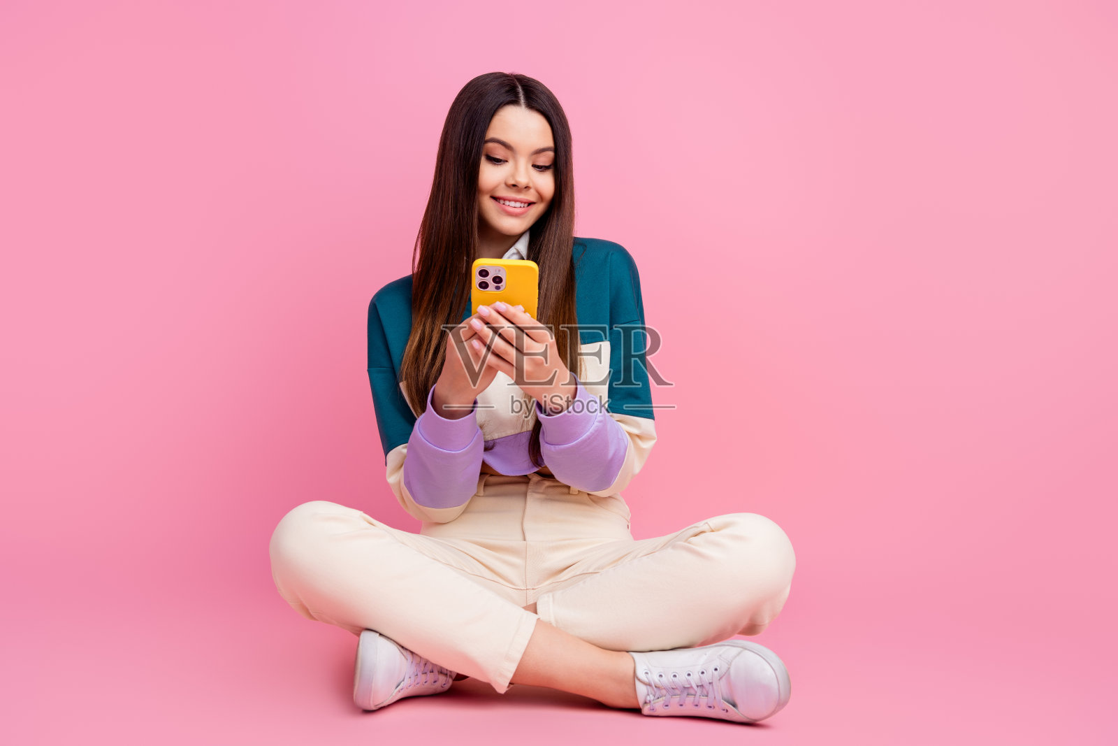 迷人的年轻快乐女性穿着休闲服装，坐在粉色背景前使用智能手机照片摄影图片