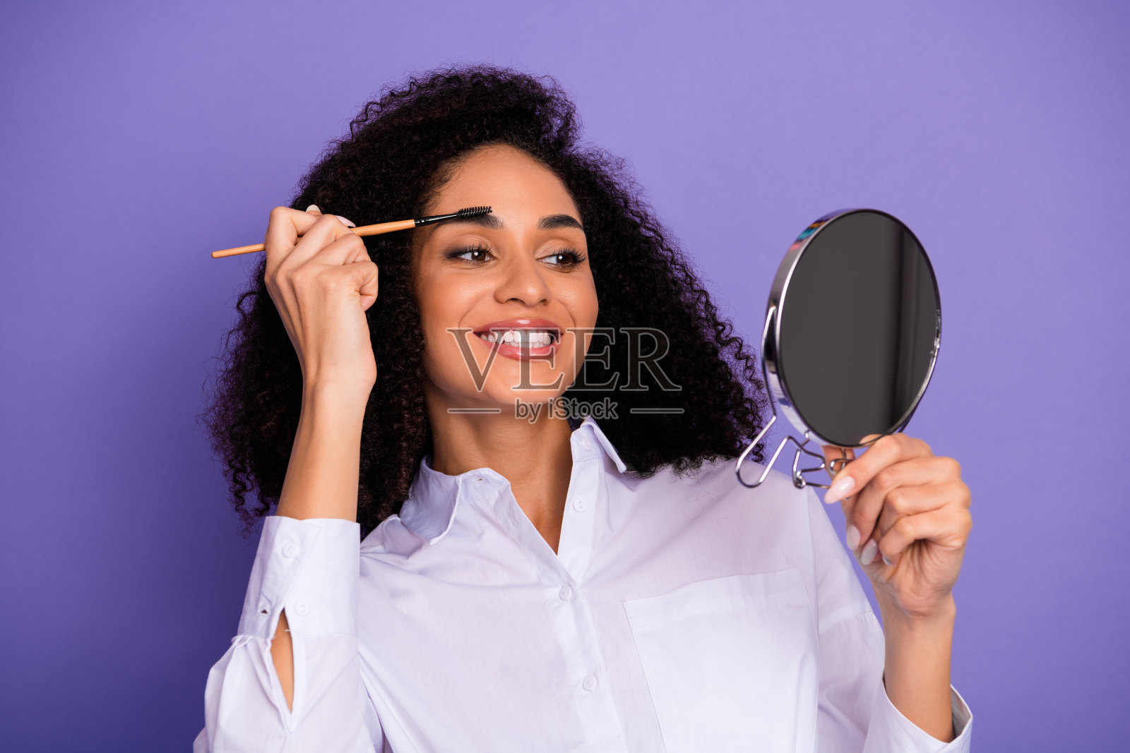 年轻女商人在紫色背景前微笑着对着镜子用刷子化妆眉毛。照片摄影图片