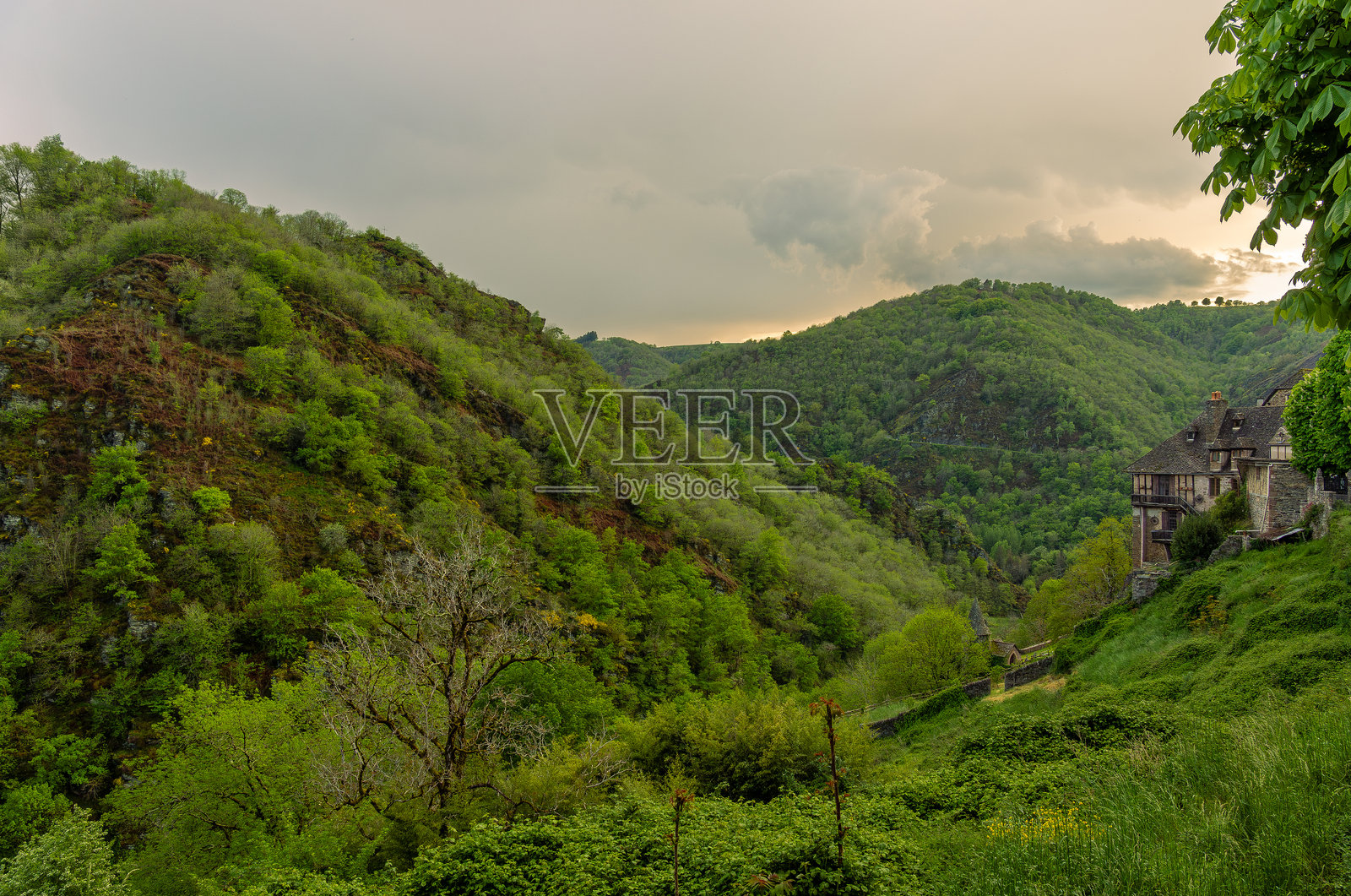 在多云的天空下，阳光透过云层洒在中世纪村庄孔克斯的绿色山脉和一座历史悠久的石屋上。照片摄影图片