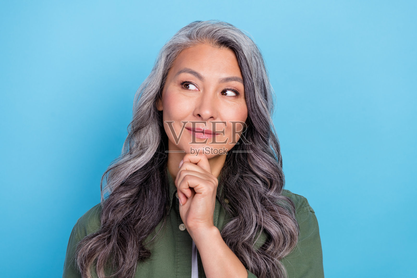 迷人而深思的退休女性，穿着绿色衬衫，手托下巴，微笑着望向空旷的空间，背景为蓝色。照片摄影图片