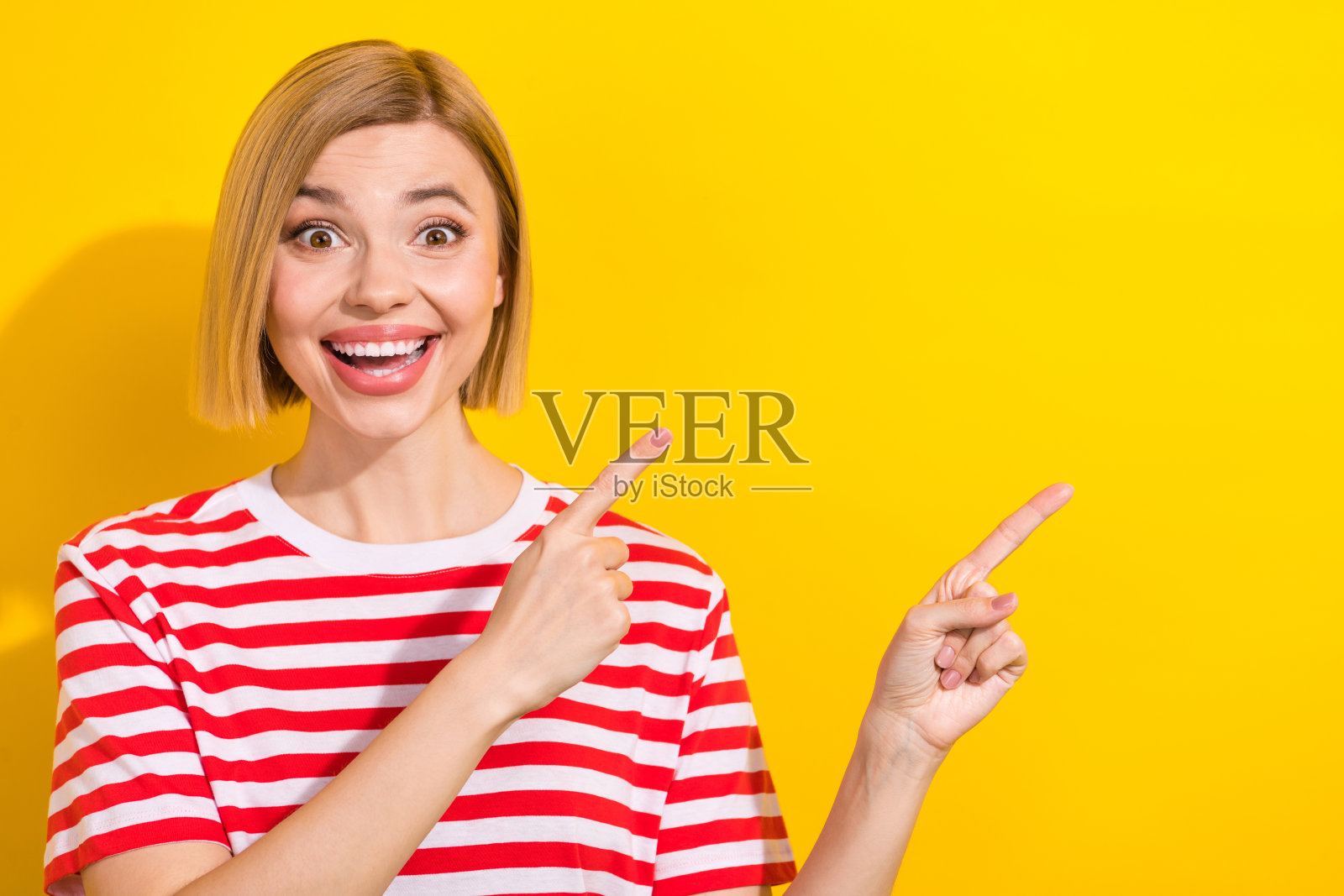 一位留着波波头、穿着条纹T恤的惊讶女性，指向令人惊叹的促销空白区域，背景为鲜艳的黄色。照片摄影图片